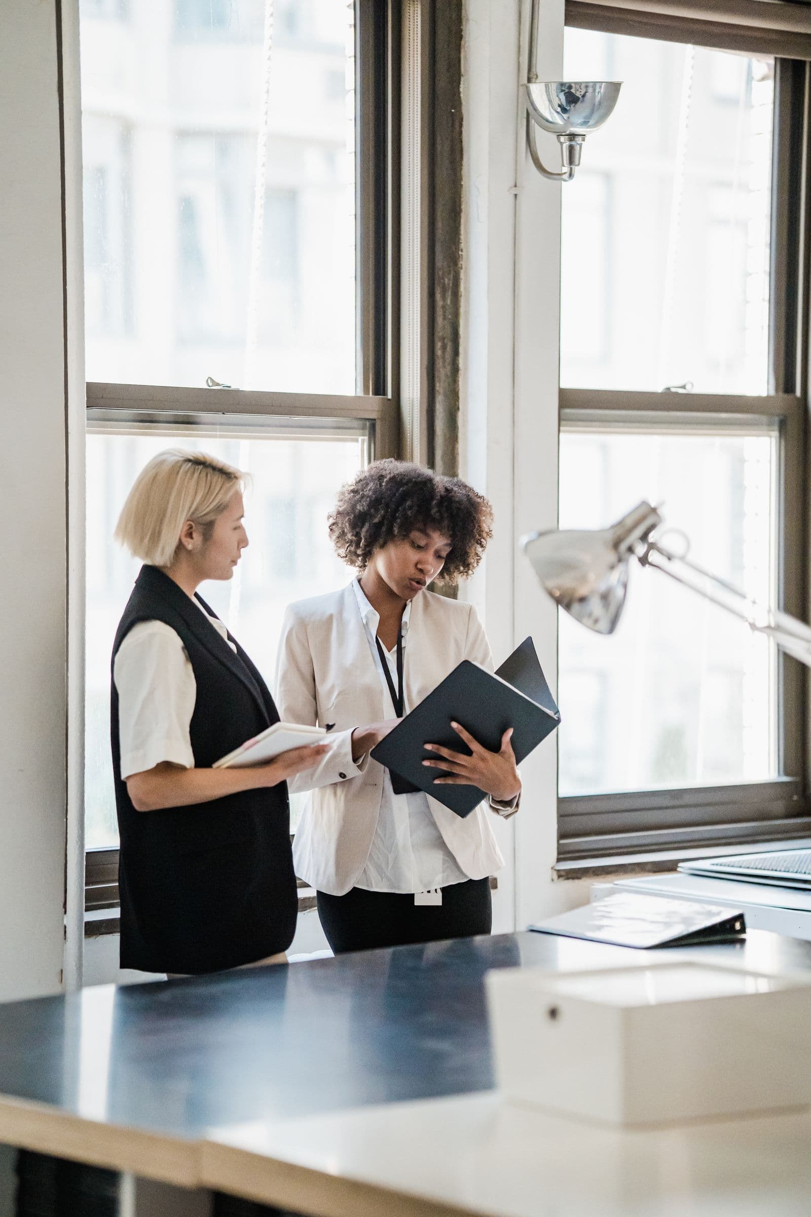 Two women reviewing an open folder and papers beside an office window